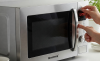 Micro-ondas moderno em uma cozinha, com painel digital visível e as mãos de uma pessoa ajustando o relógio. Ao lado, recipientes seguros para microondas, como um pirex de vidro e um prato de cerâmica, estão organizados sobre a bancada. A imagem transmite praticidade, organização e uso correto do eletrodoméstico